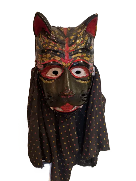 Handcrafted mask resembling a cat with wide eyes, red and gold painted details, and a black cloth with multicolored polka dots draped beneath.