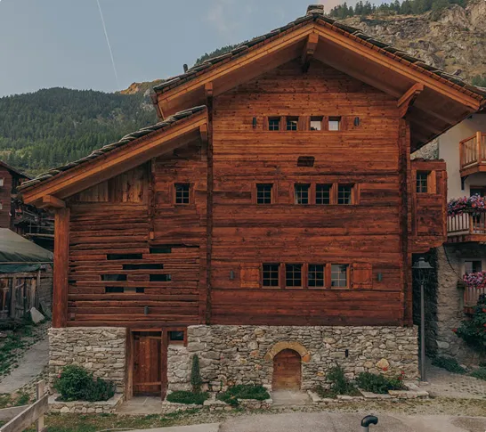 Traditional wooden chalet with stone foundation set against forested mountain backdrop.