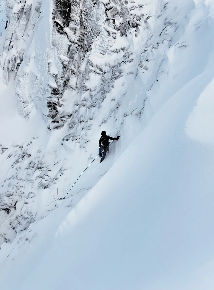 Person ice climbing on a steep, snowy mountain slope with rugged, snow-covered rocks.