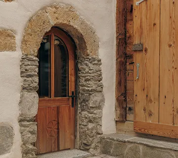 Small arched wooden door set in rough stone and plaster wall next to a larger wooden door.