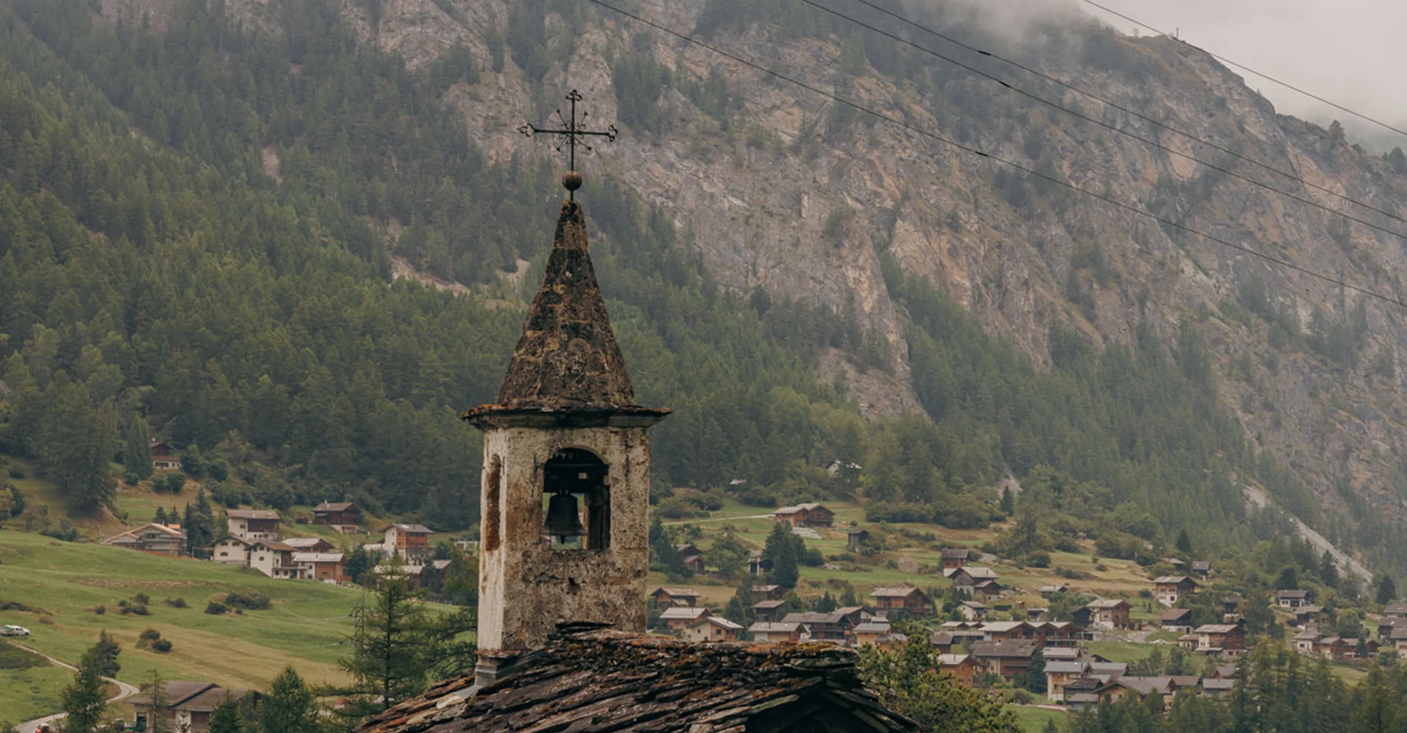 Rustic church bell tower with a cross overlooking a small village and forested mountain in the background.