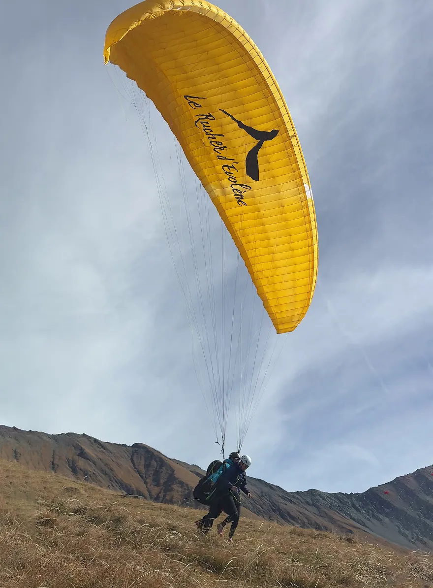 Paraglider with a bright yellow canopy taking off from a grassy hillside with mountains in the background.