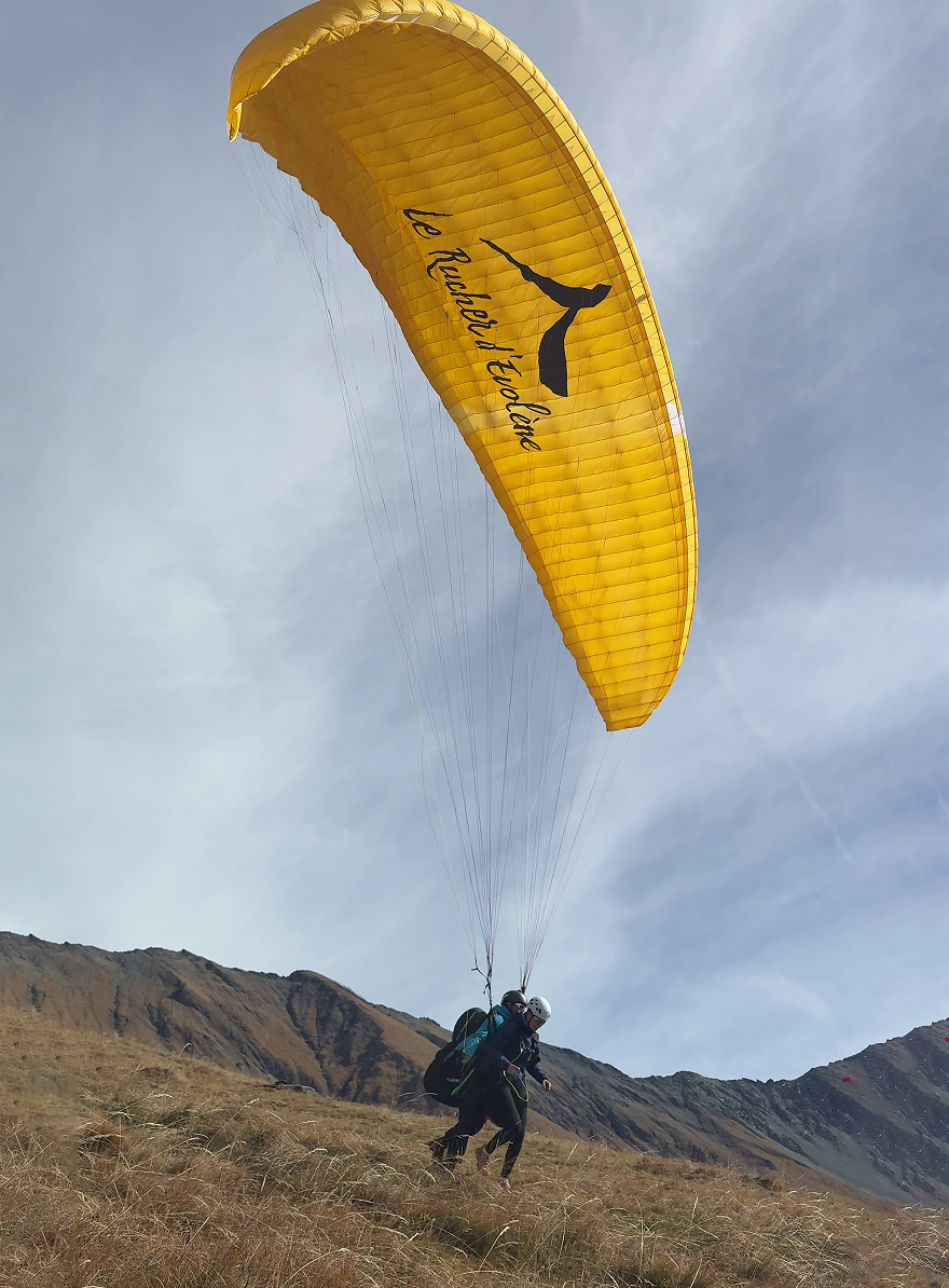 Two people preparing to paraglide with a large yellow wing above a grassy mountain slope under a cloudy sky.
