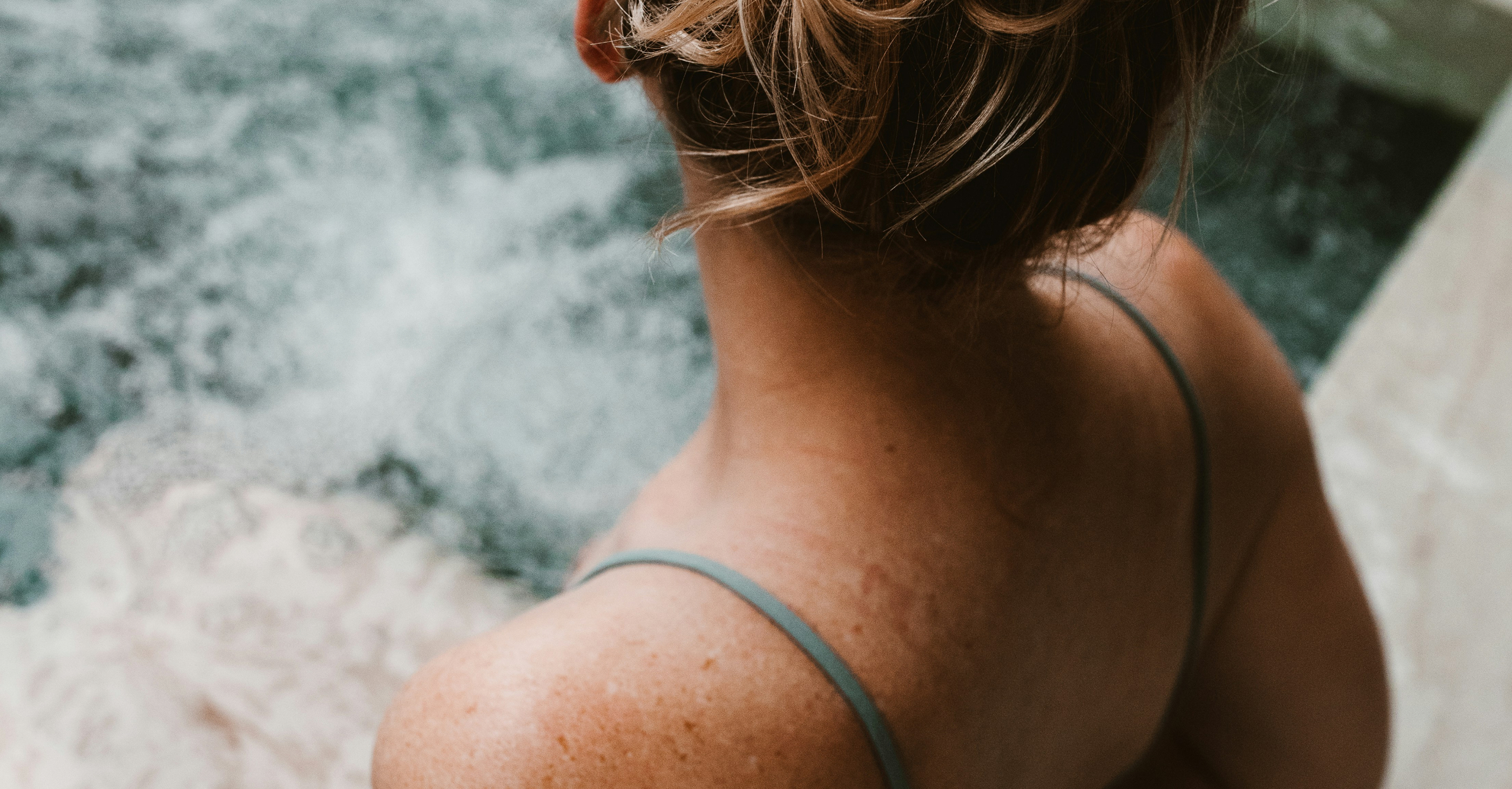 Woman with light brown hair tied up wearing a gray strap top, sitting near bubbling water, viewed from behind.
