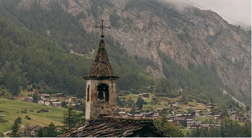 Old church tower with a cross overlooking a village in a mountainous forested area.