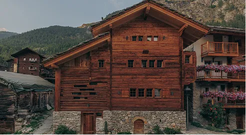 Rustic wooden chalet with stone base amid mountain landscape and nearby buildings with flower balconies.