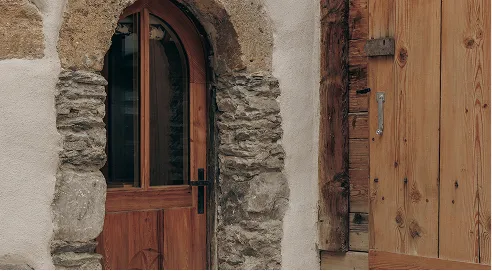 Wooden door with glass panels set in a stone archway next to a wooden shutter on a beige wall.