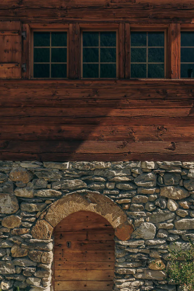 Rustic building facade with stone base, wooden arched door, and upper wooden walls with four square windows.