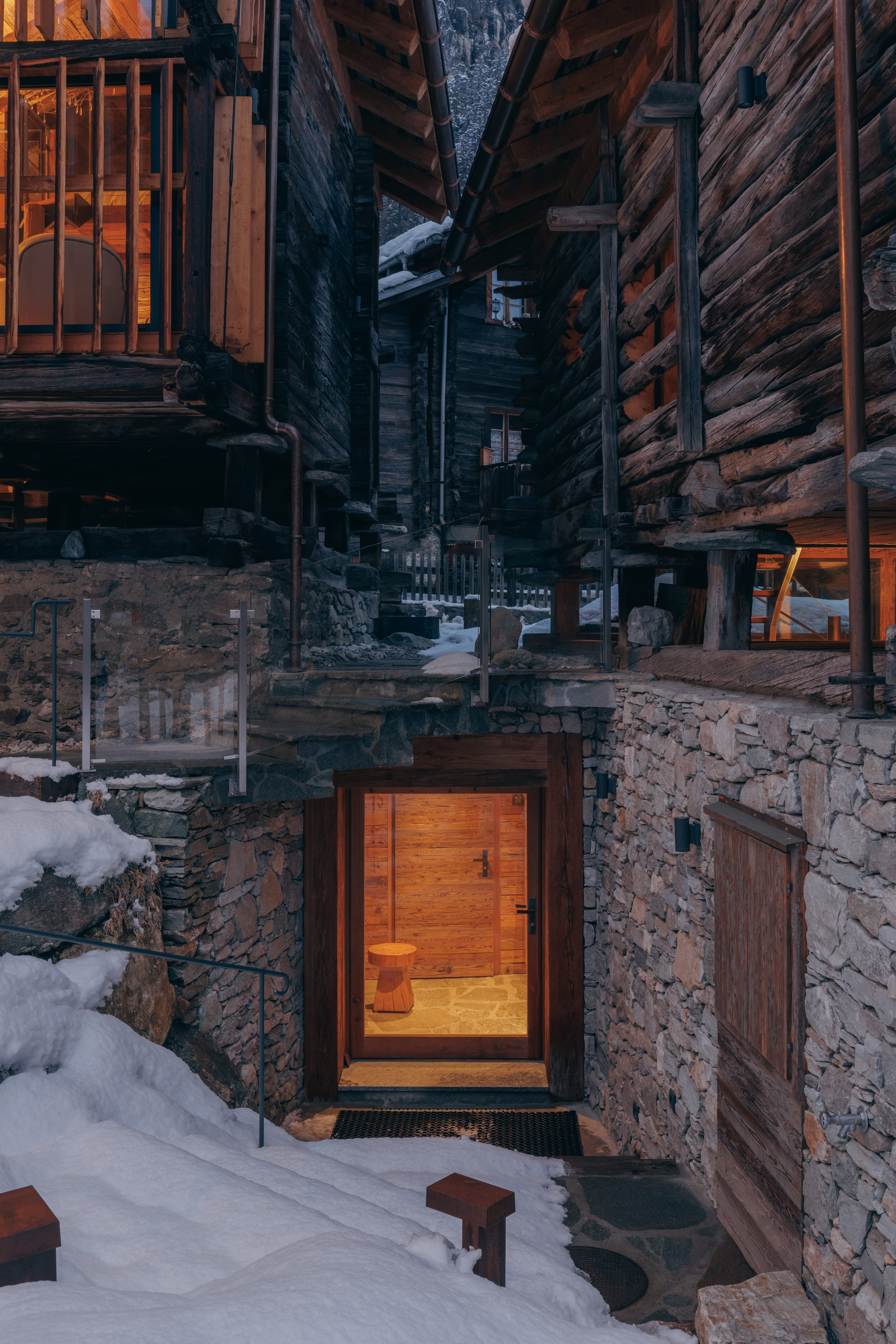 Stone and wooden cabin entrance with warm light inside, surrounded by snow and wooden buildings.
