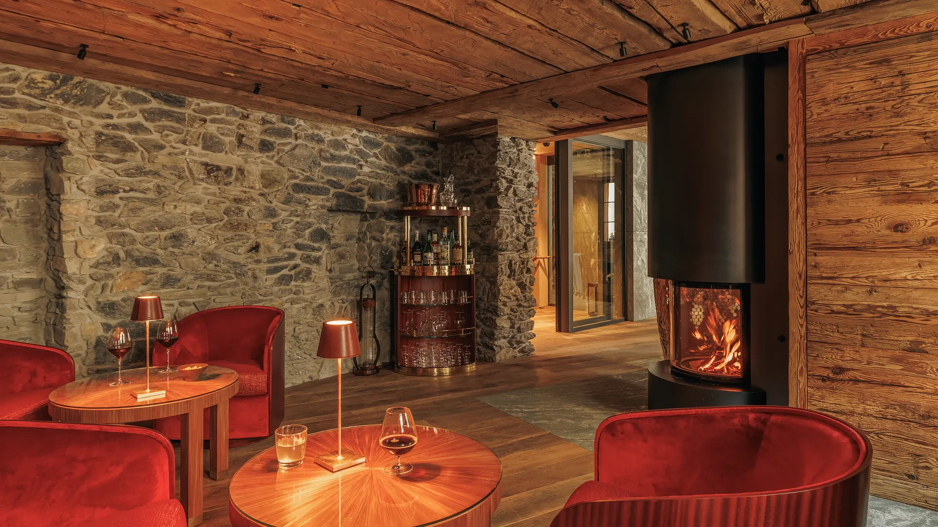 Cozy rustic lounge with red velvet armchairs around wood tables lit by small lamps, a stone wall, a modern black fireplace with fire, and a bar cart with bottles and glasses.