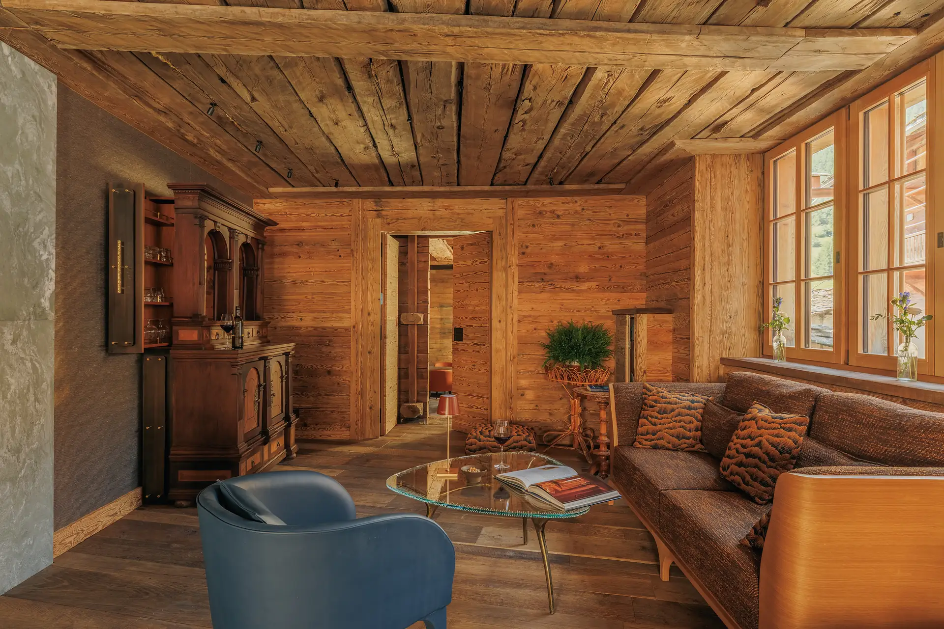 Cozy living room with wooden walls and ceiling, featuring a brown couch with patterned pillows, a blue armchair, a glass-top coffee table with a book and a glass of wine, and a wooden cabinet with glassware, next to large windows with flower vases.