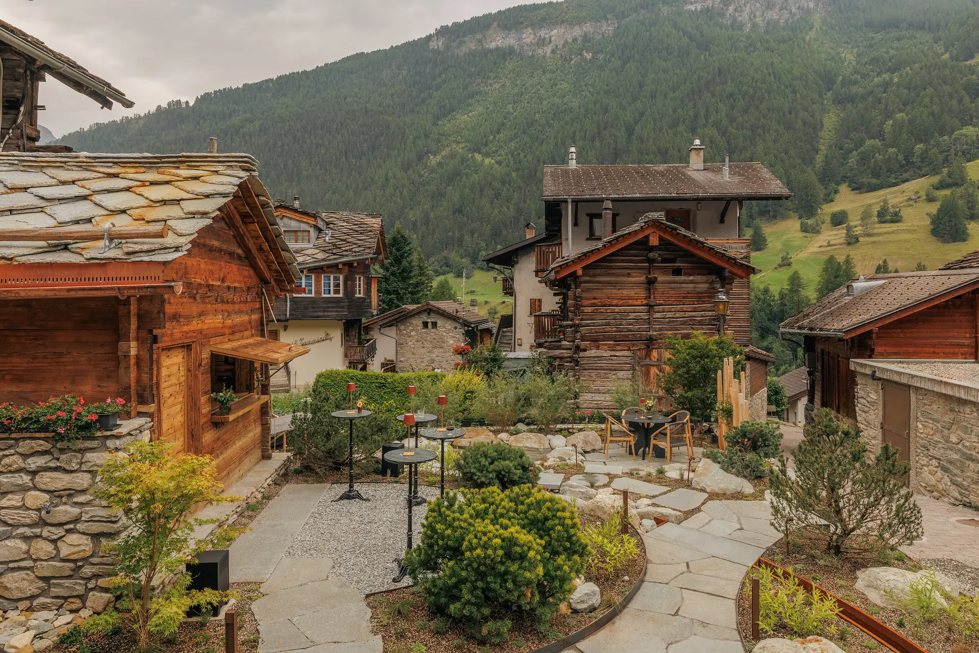 Outdoor seating area with small round tables and chairs surrounded by lush greenery and stone paths, set among rustic wooden and stone mountain houses with forested hills in the background under a cloudy sky.