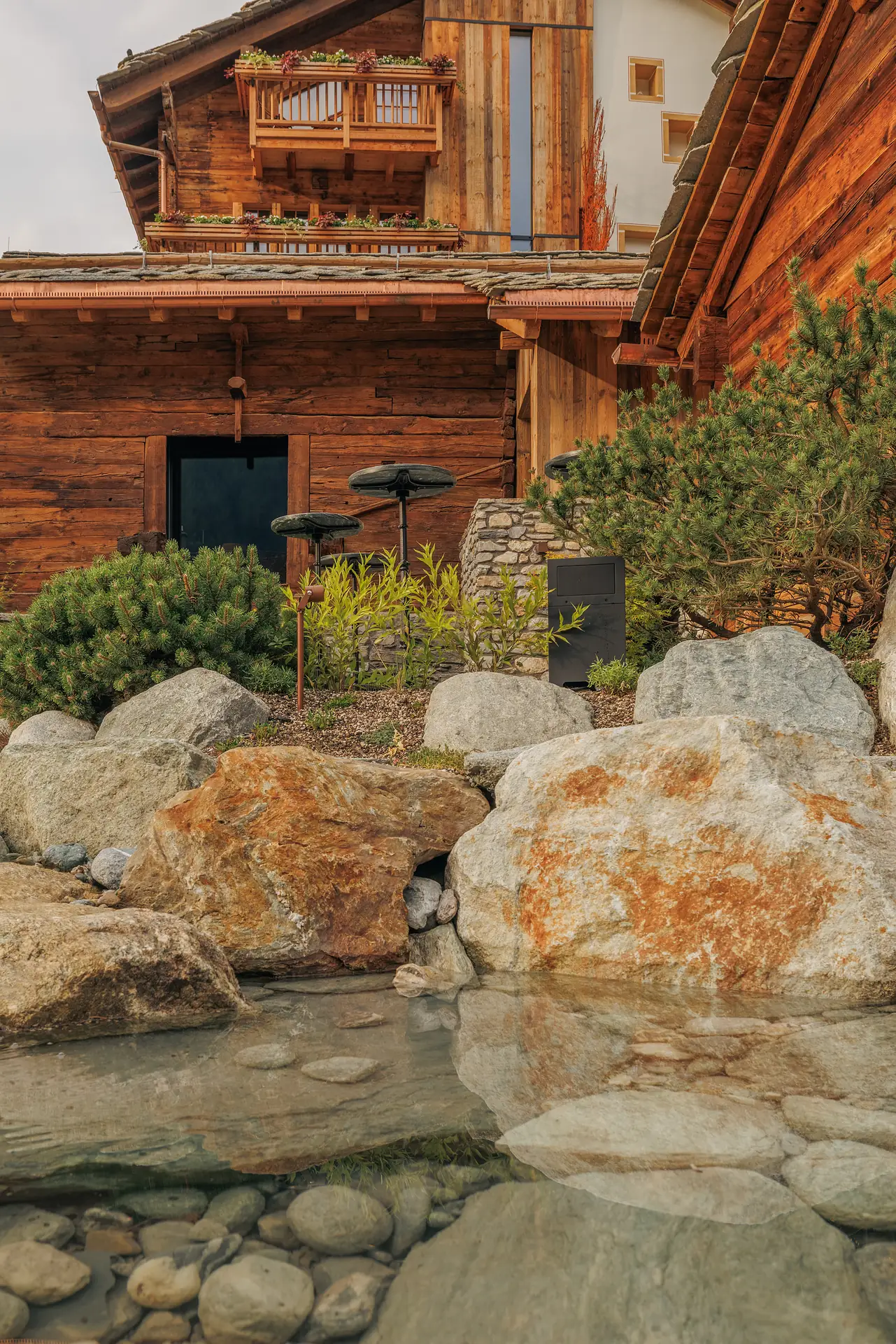 Rustic wooden chalet with a balcony adorned with flower pots above, surrounded by large natural stones, pine bushes, and a clear shallow pond reflecting the scene.