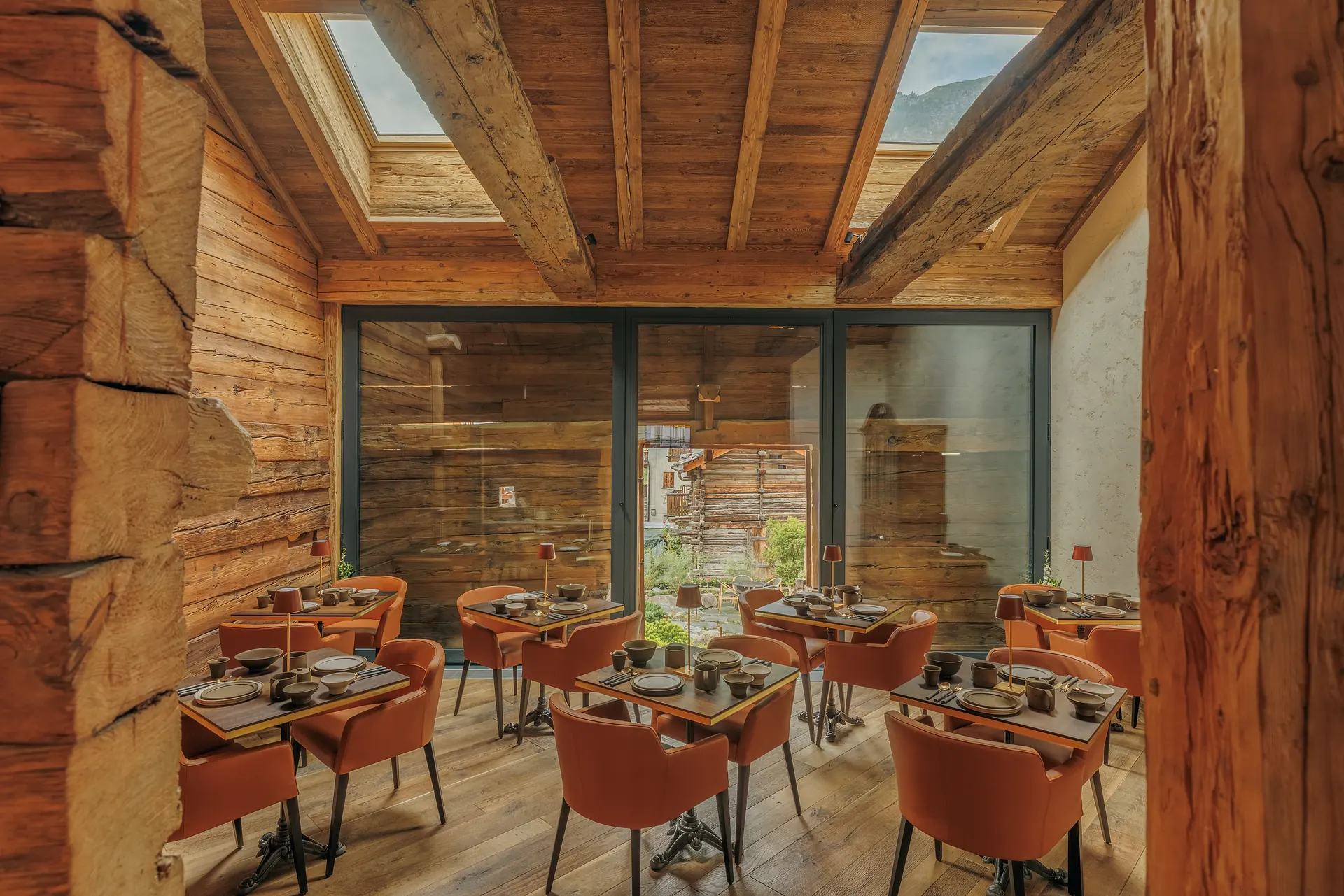 Cozy indoor dining room with wood-paneled walls and ceiling, exposed wooden beams, six square tables set for meals with brown chairs, and large glass doors showing an outdoor view.