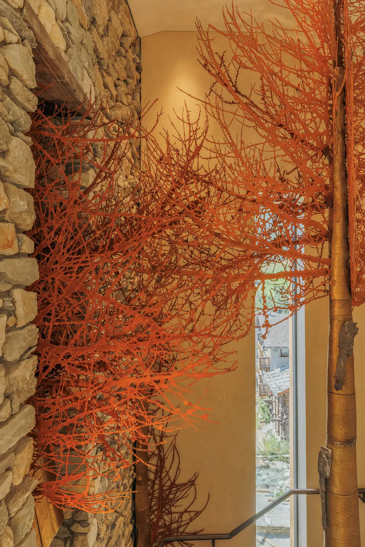 Interior staircase with stone walls and decorative branches painted bright orange mounted on the wall and ceiling, next to a tall window showing a view of a building outside.