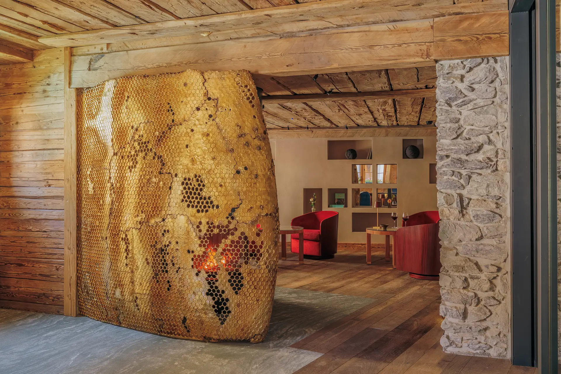 Interior of a cozy rustic room featuring a unique honeycomb-patterned fireplace with a fire burning inside, wooden walls and ceiling, stone column, and two red armchairs around wooden tables with glasses of red wine.