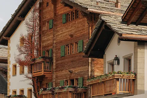 Wooden and stone alpine chalet with green window shutters and small balconies decorated with flowers, under a cloudy sky.