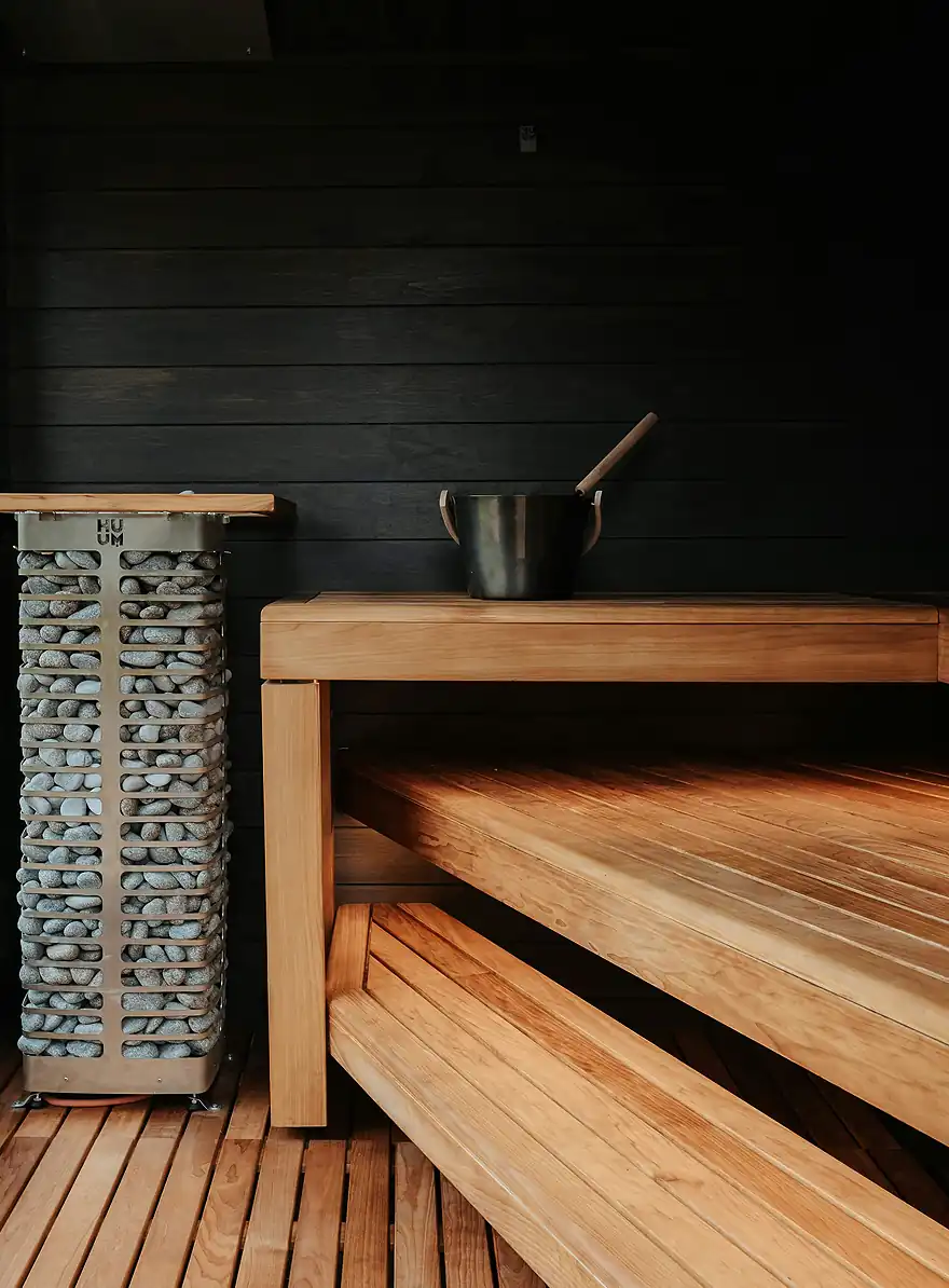 L'intérieur moderne du sauna comprend un poêle en métal rempli de pierres rondes, un banc en bois avec un seau noir et une louche sur le dessus, le tout contre un mur lambrissé en bois foncé.