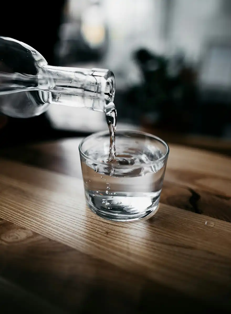 De l'eau est versée d'une bouteille transparente dans un petit verre transparent posé sur une surface en bois.