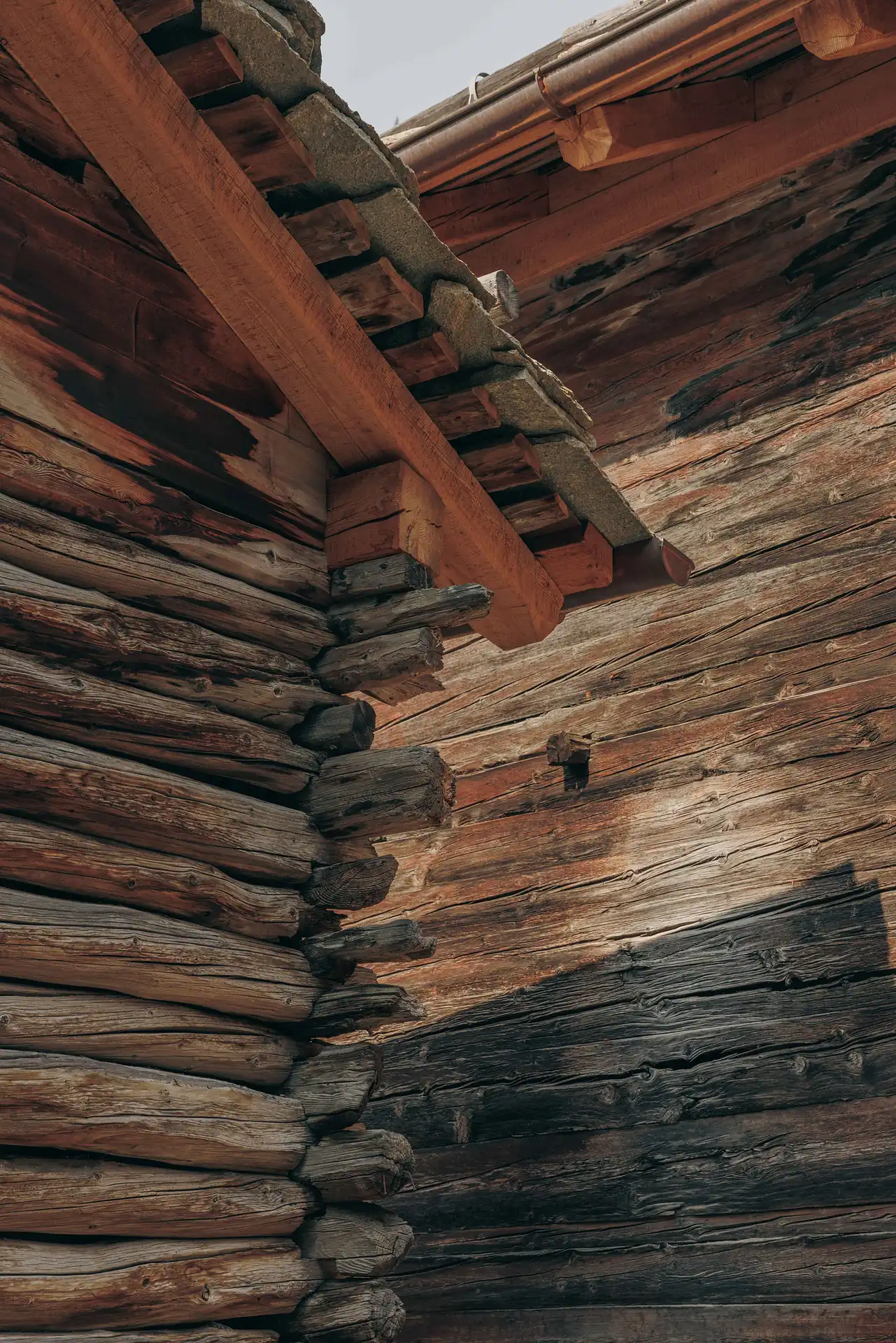 Close-up view of corner and eave of a rustic log cabin roof showing weathered wooden logs and wooden beams supporting the stone-covered roof edge.