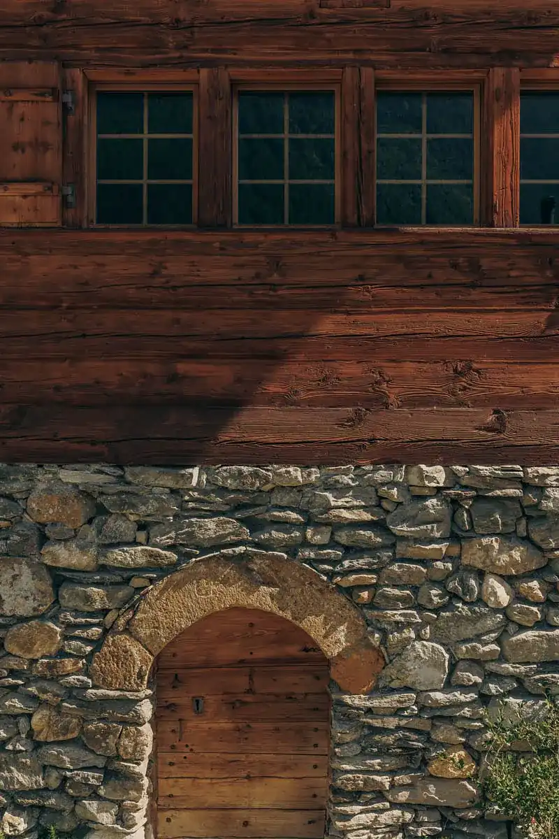 Facade of a rustic building with a lower section made of rough stone featuring an arched wooden door, and an upper section of wooden planks with four small grid-style windows.