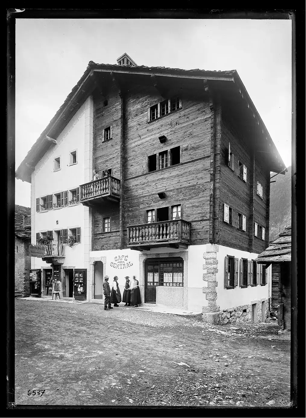 Black and white photo of a large traditional chalet building with wooden and plaster exterior walls, two balconies, and multiple shuttered windows; four people in period clothing stand near the entrance labeled 'Cafe Central,' and two women look out from a balcony on the upper floor; the ground in front is dirt with some scattered stones.