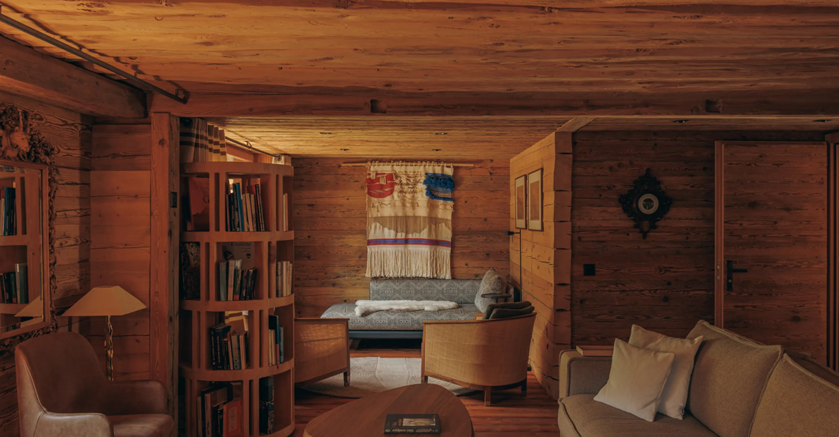Cozy wooden living room with a gray sofa, two wooden armchairs, a round coffee table, a vertical wooden bookshelf with books, a tall lamp, a decorative wall hanging, and warm lighting.