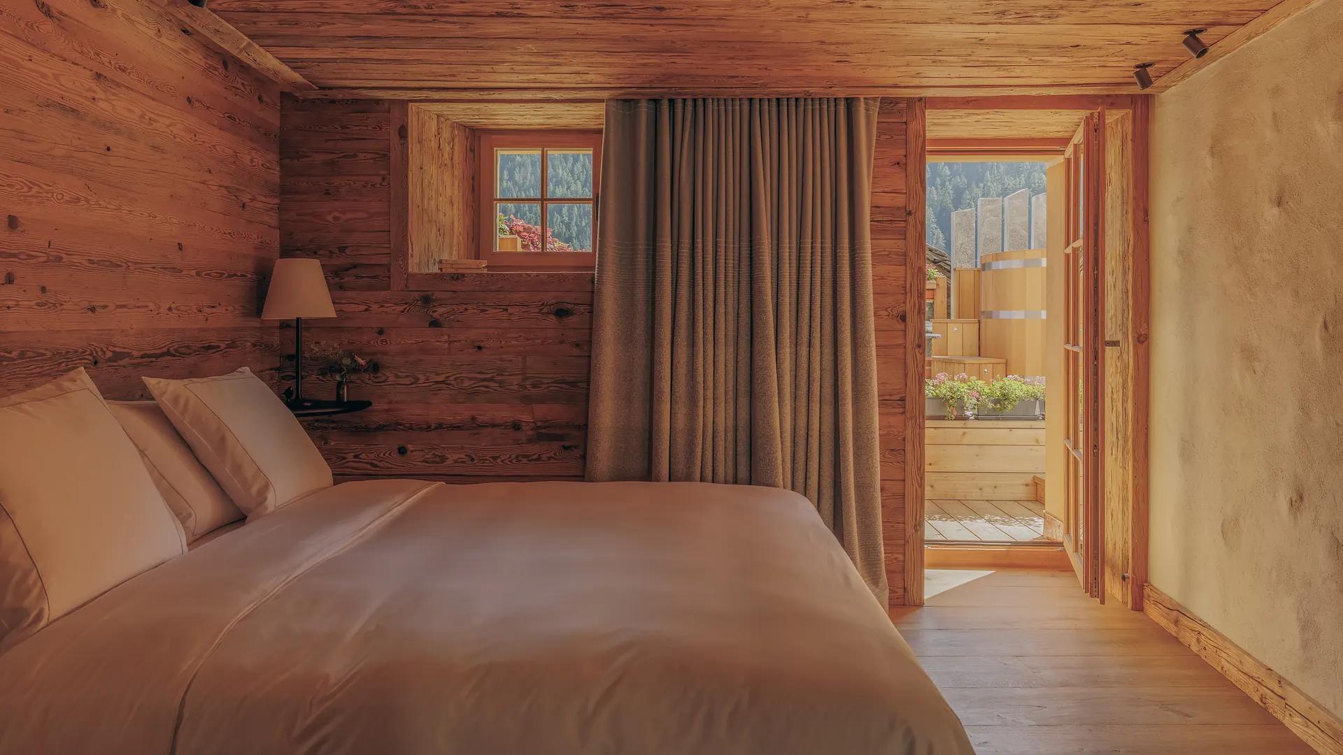 Bed with linen pillows in a log-walled room, door open onto a private terrace with a wooden hot tub