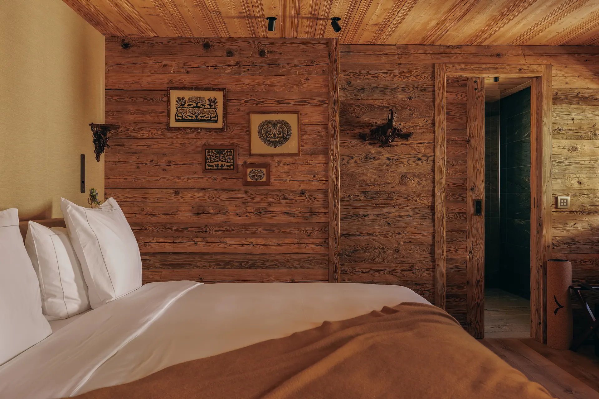 Close-up of a double bed with white linen and a camel throw, framed folk art prints on the log wall beside it