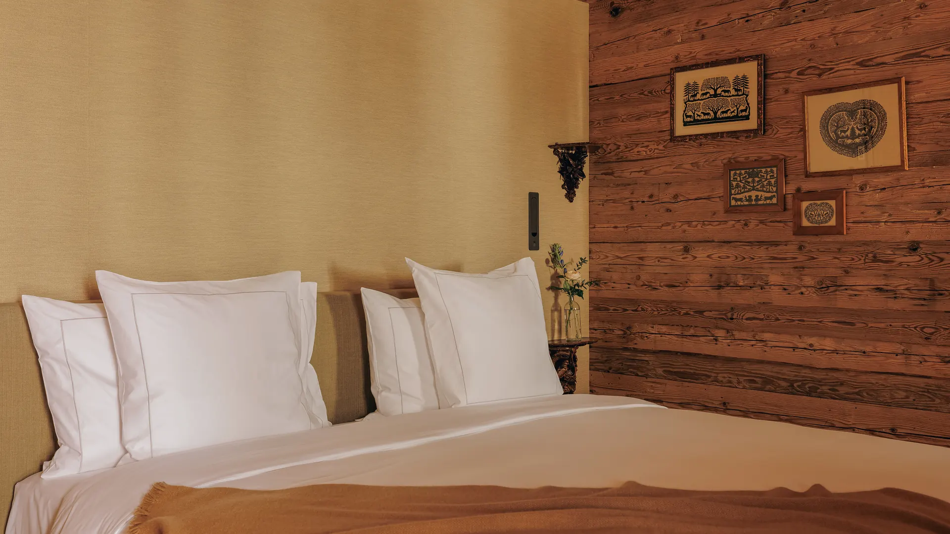 Wide-angle view of the bed headboard area, four folk art prints on the log wall and camel throw across white bedding