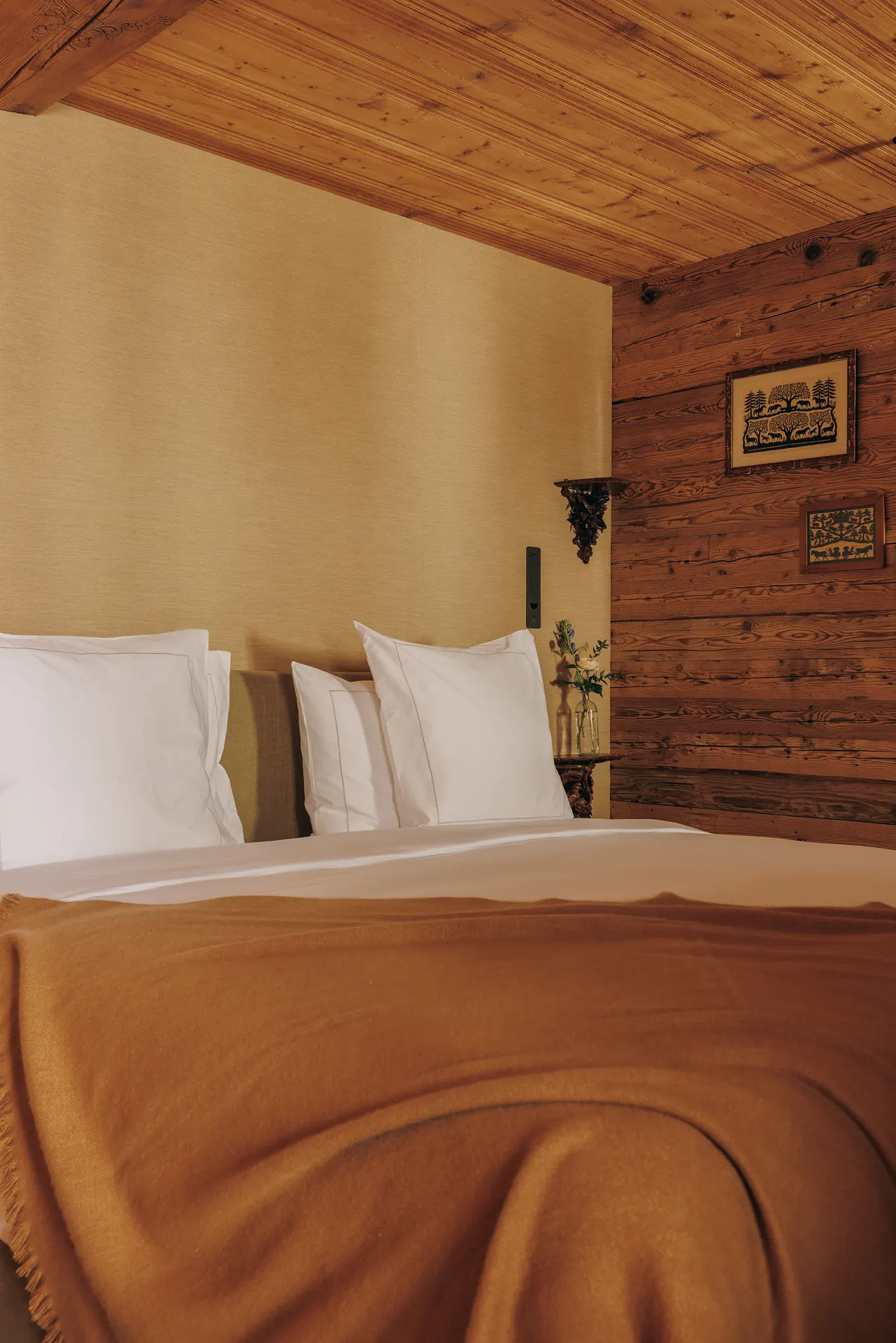 Wide-angle view of the bed headboard area, four folk art prints on the log wall and camel throw across white bedding