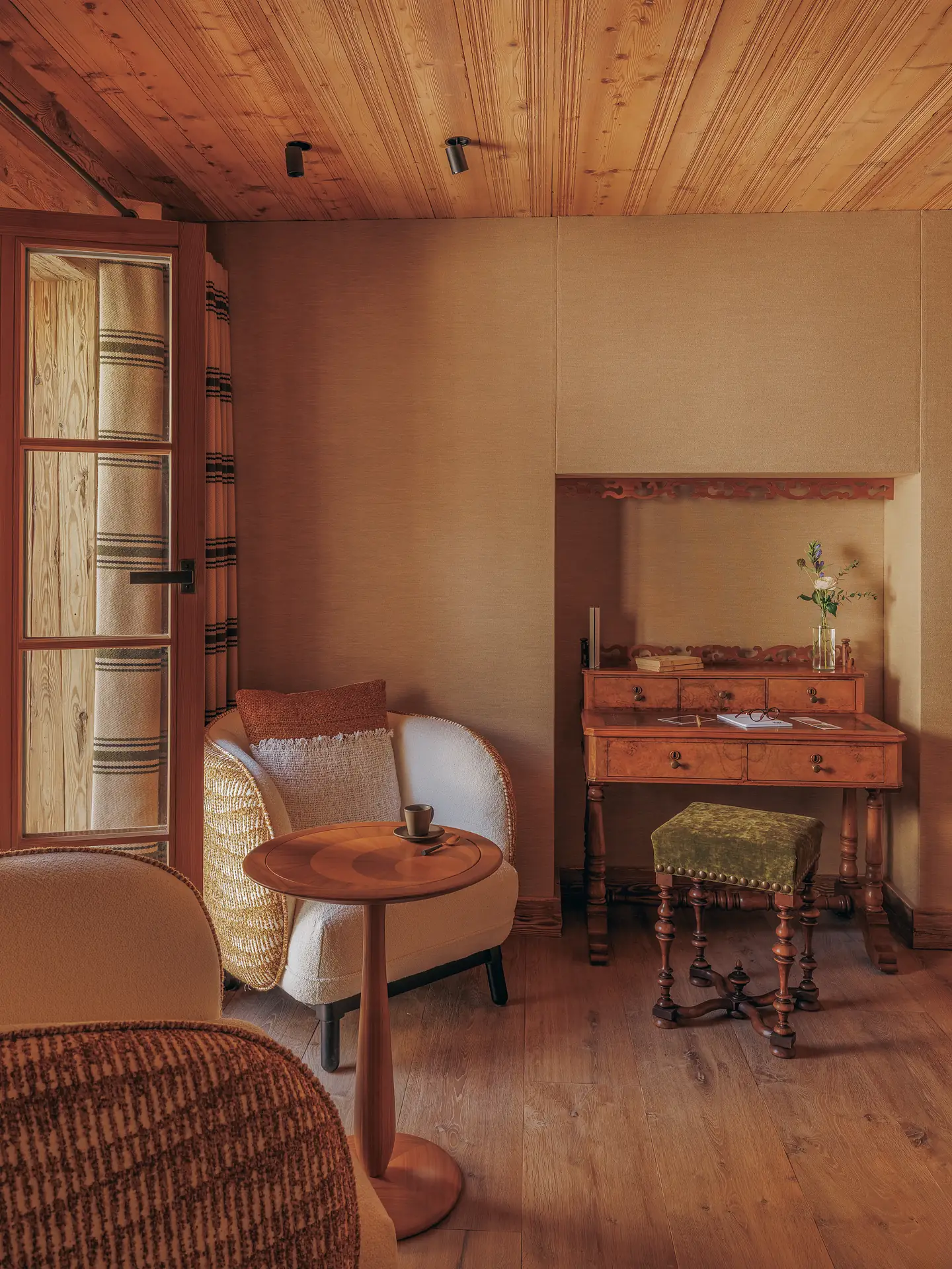 Sitting area with two armchairs and a round side table, antique writing desk and green velvet stool in a beige-panelled alcove