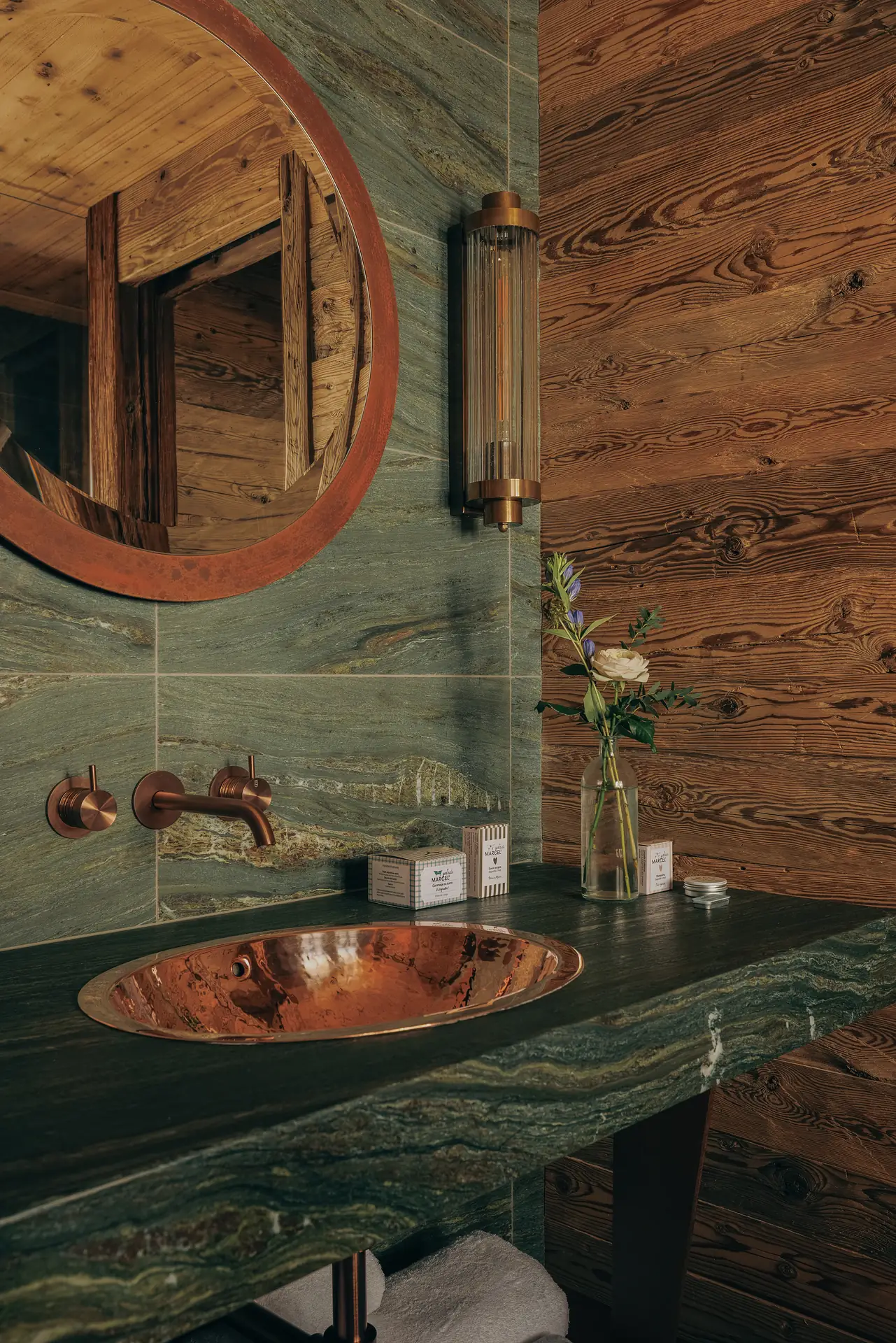 Oval copper basin set in a green marble countertop, with a wall-mounted brass tap, round copper mirror and a small flower vase