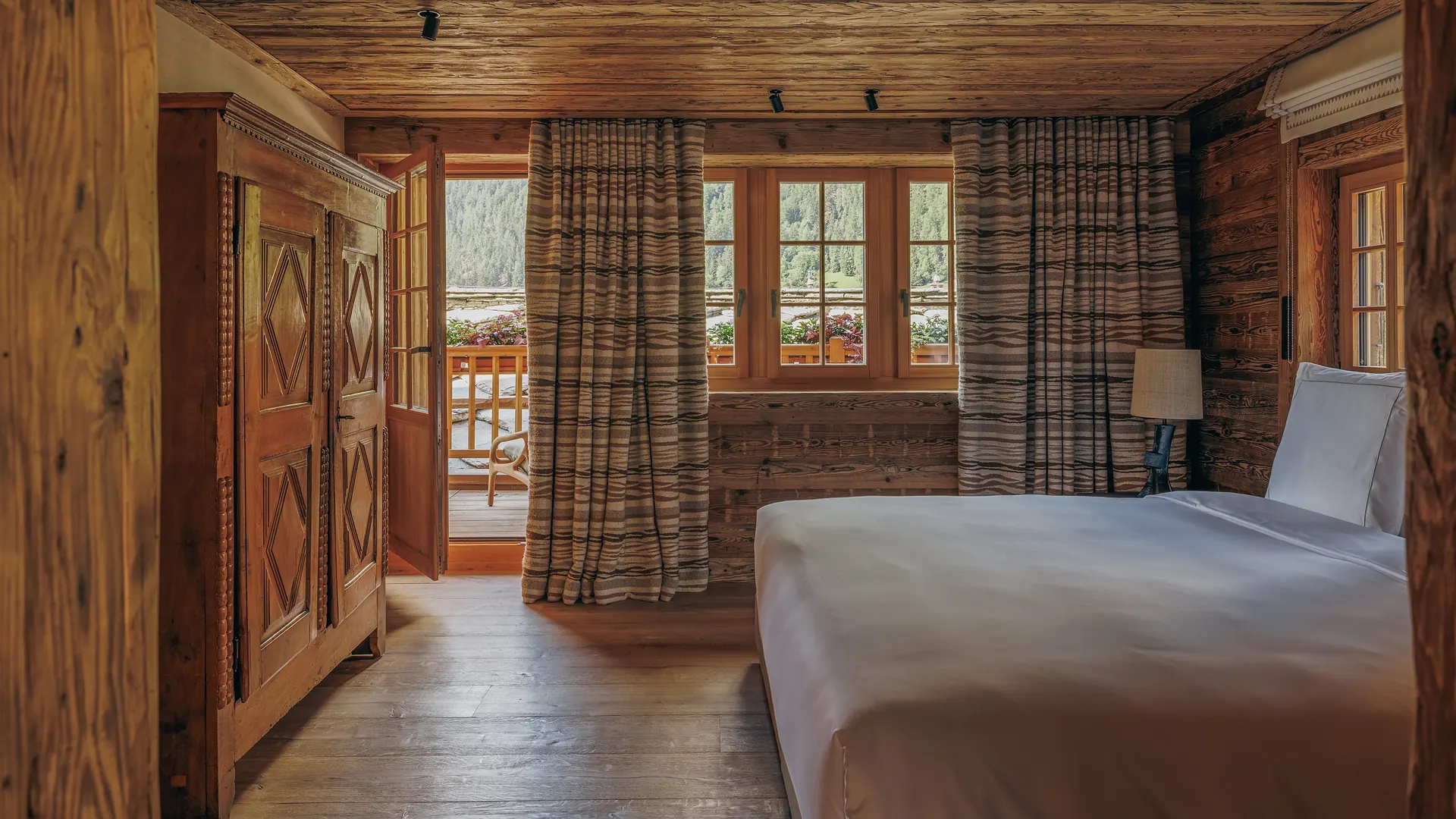 Bedroom with a carved alpine wardrobe, balcony door open onto a flower-lined terrace, striped curtains and a double bed
