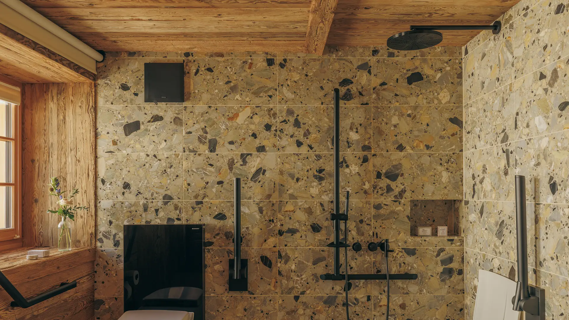 Duschwand gefliest mit großformatigem Terrazzo, dazu eine Regendusche und Armaturen in Mattschwarz, links ein Fenster mit Blumen