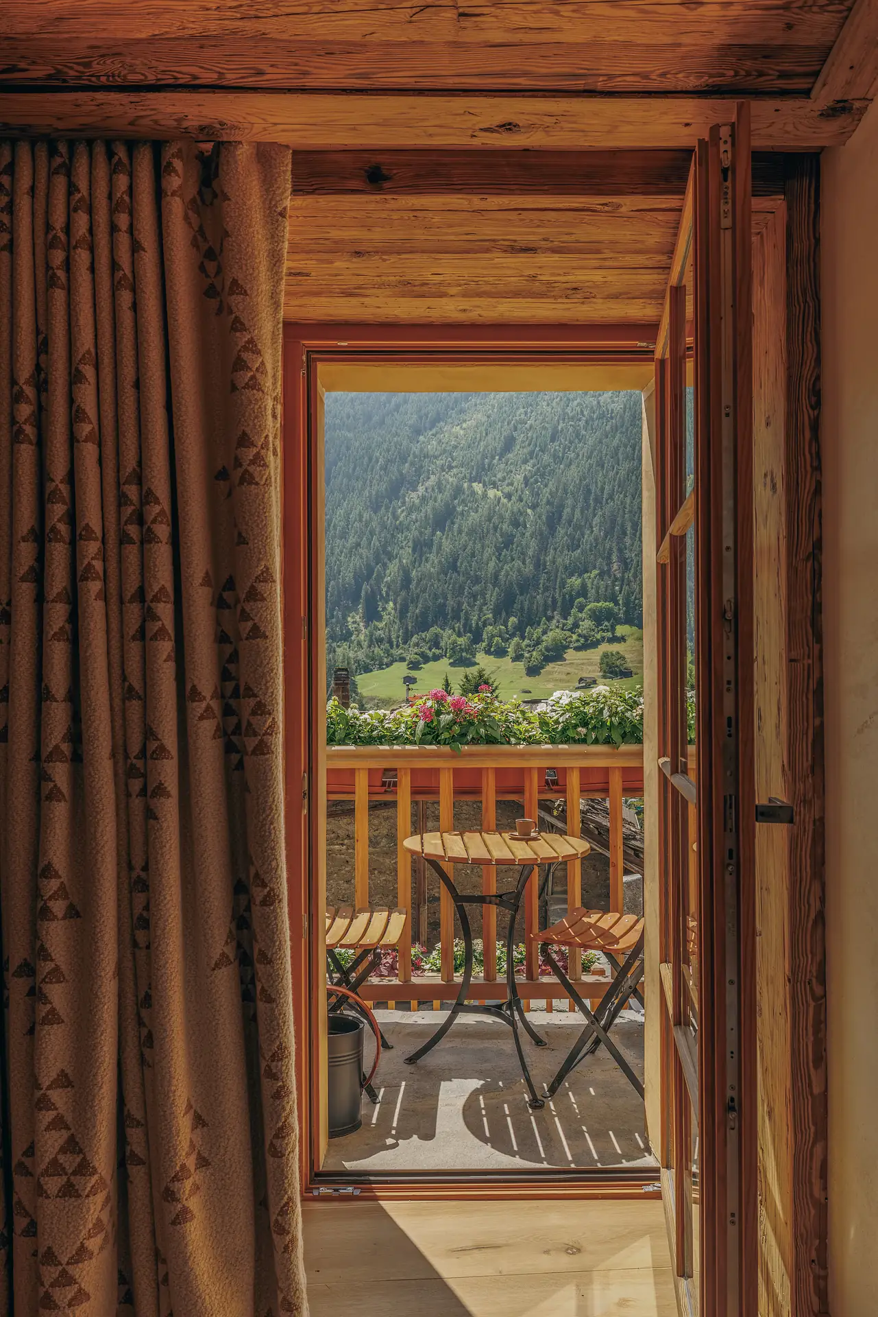 Blick auf einen Balkon durch eine offene Tür, eingerahmt von Vorhängen mit Dreiecksmuster; auf dem Balkon stehen ein Bistrotisch und Stühle mit Aussicht auf einen Kiefernwald