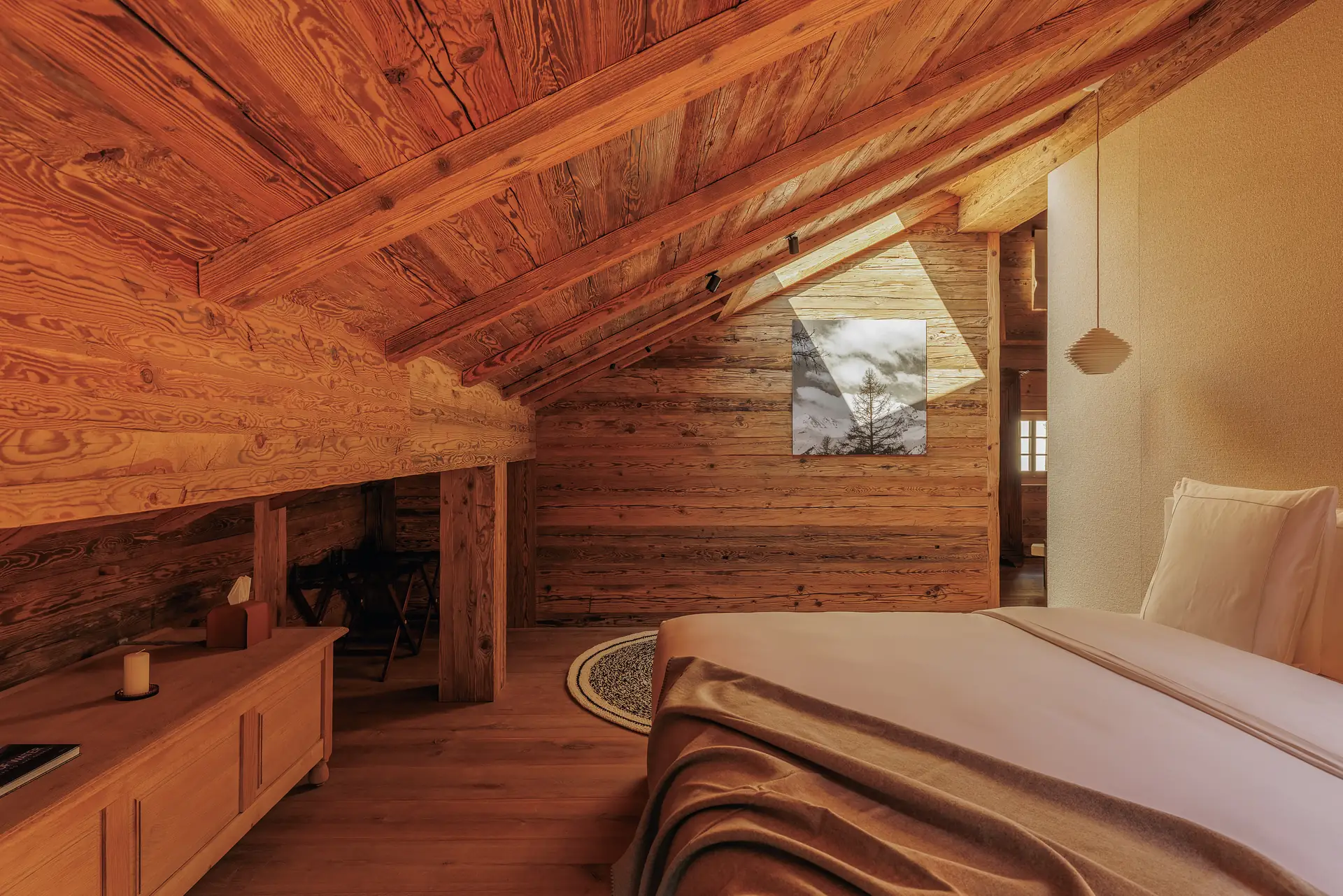 Attic bedroom with exposed log beams, a double bed, a Le Corbusier chaise longue and a balcony door open onto the valley