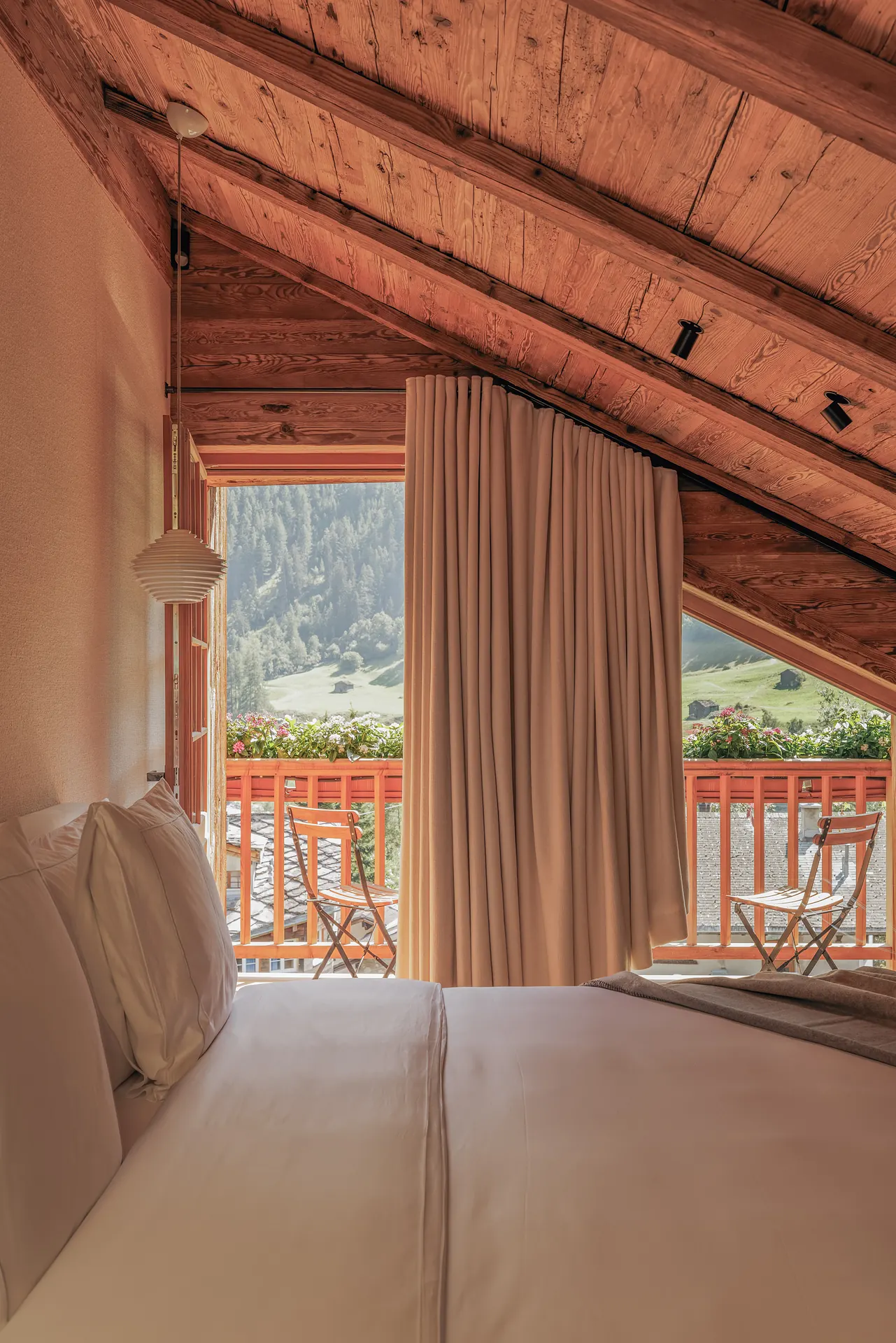 Bed with white linen below a sloped log ceiling, balcony door open to a mountain view and flower-lined railing