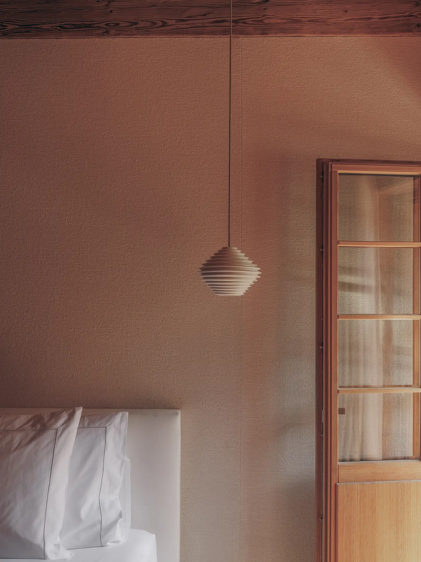 Bedroom detail showing a ribbed pendant lamp hanging above white pillows against a textured terracotta wall