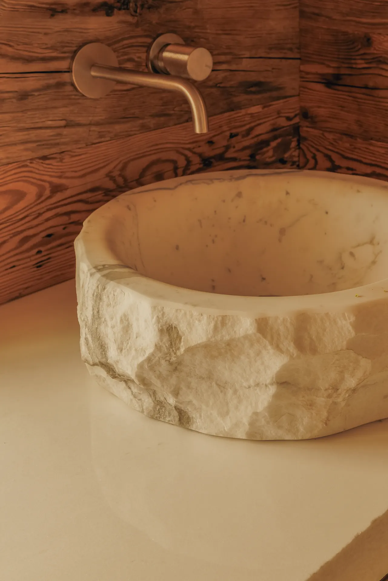 Close-up of a hand-carved white marble vessel basin with a rough-hewn exterior and a wall-mounted brass tap