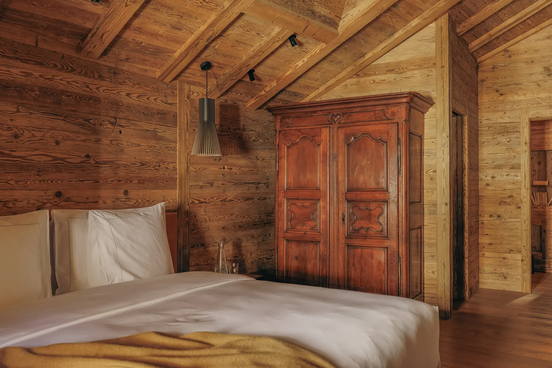 Schlafzimmer Etheila mit Doppelbett, Lamellen-Pendelleuchte und antikem Schnitzschrank vor Blockwänden unter einer Dachschräge