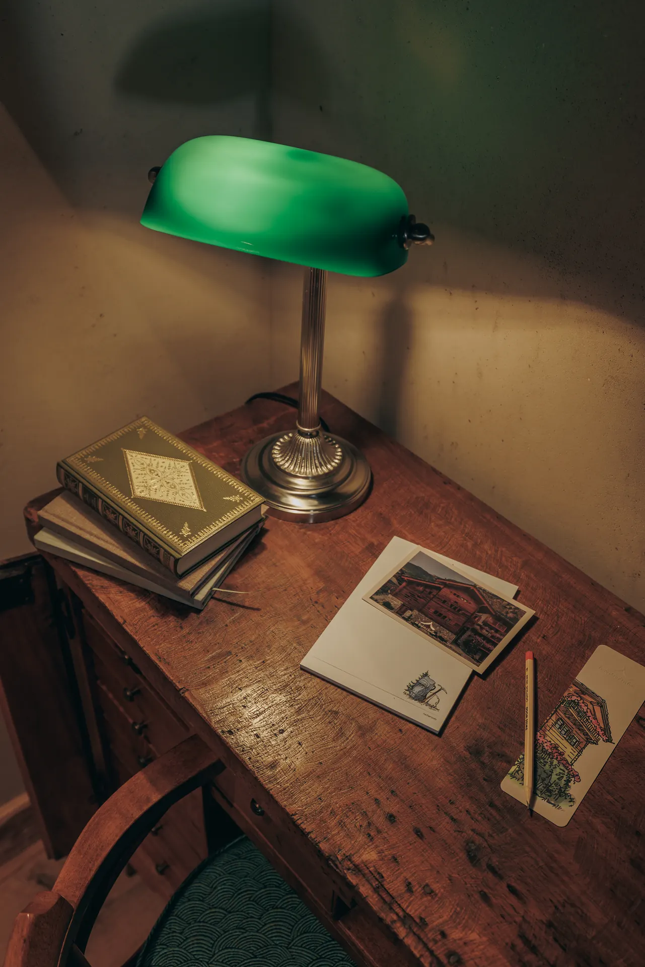 Antiker Holzschreibtisch mit grüner Bankerlampe, Bücherstapeln sowie Hotelpostkarten und Briefpapier