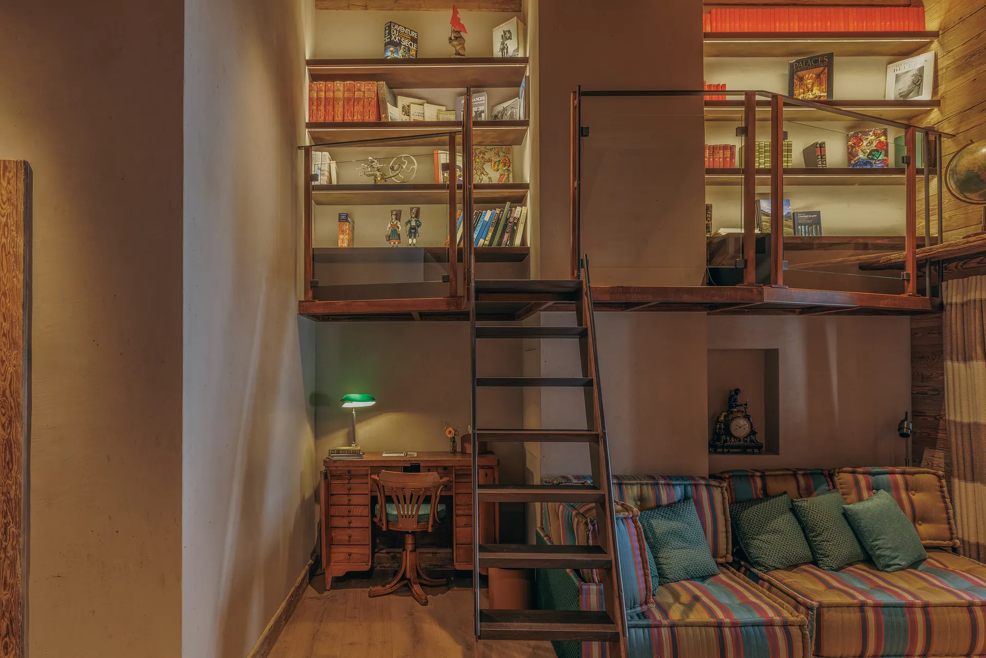 Bibliotheks-Mezzanin mit beleuchteten Regalen, einer Leiter, einem farbenfroh gestreiften Sofa und einem antiken Schreibtisch darunter