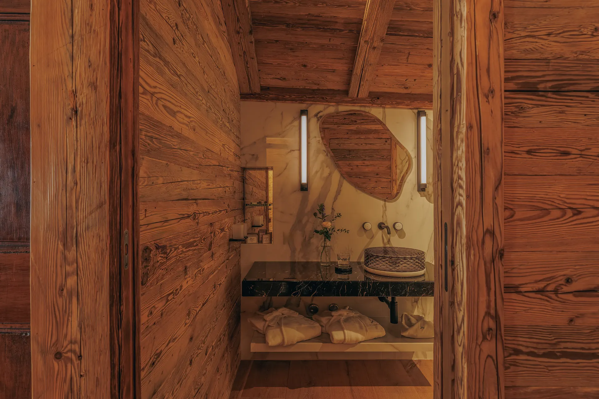Blick durch die Holztüren auf das Badezimmer mit organisch geformtem Spiegel, Waschtisch aus schwarzem Marmor, Aufsatzwaschbecken und gefalteten Bademänteln auf dem Boden