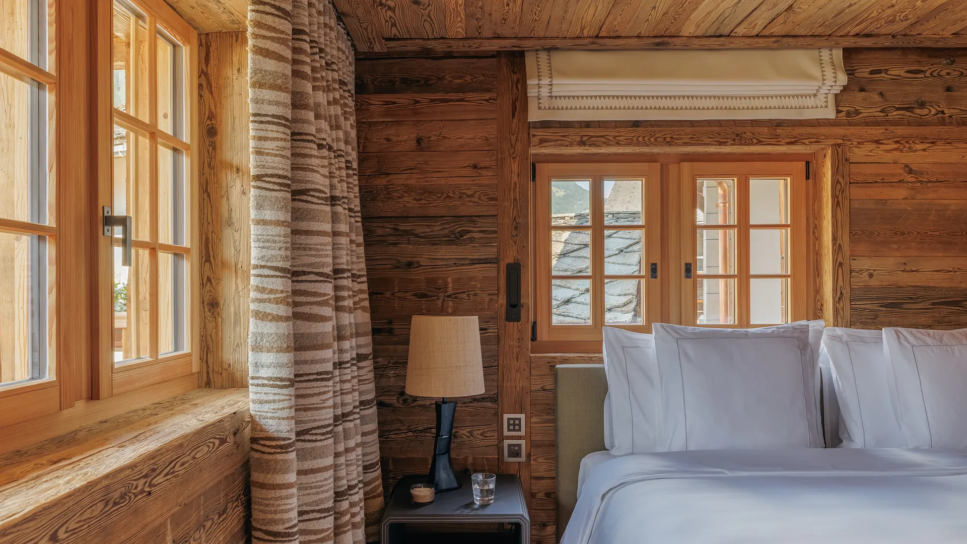 Bedside table with lamp beside striped curtains and windows overlooking rooftops