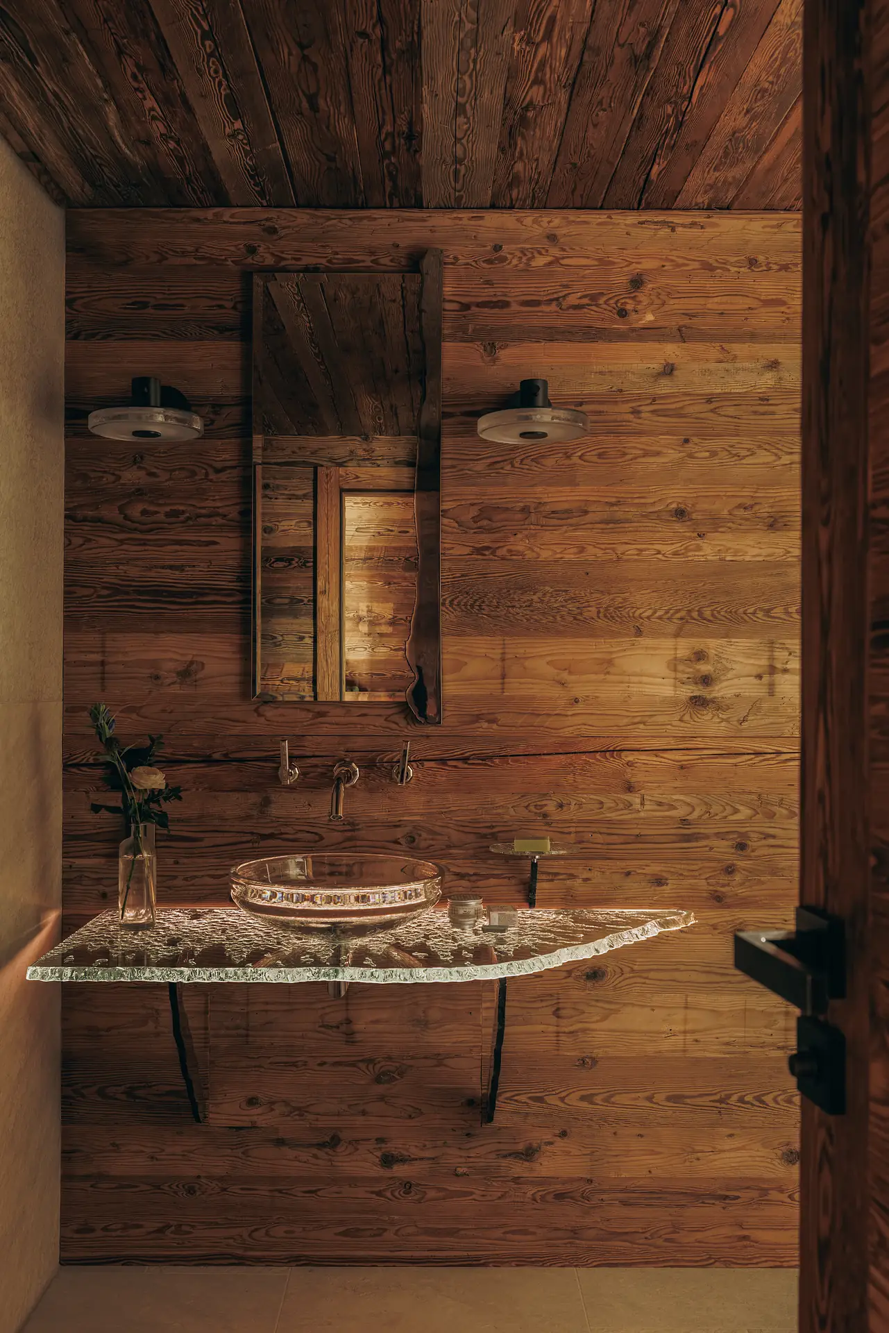 Wall-mounted faucet above a glass vessel sink with a mirror and fresh flowers