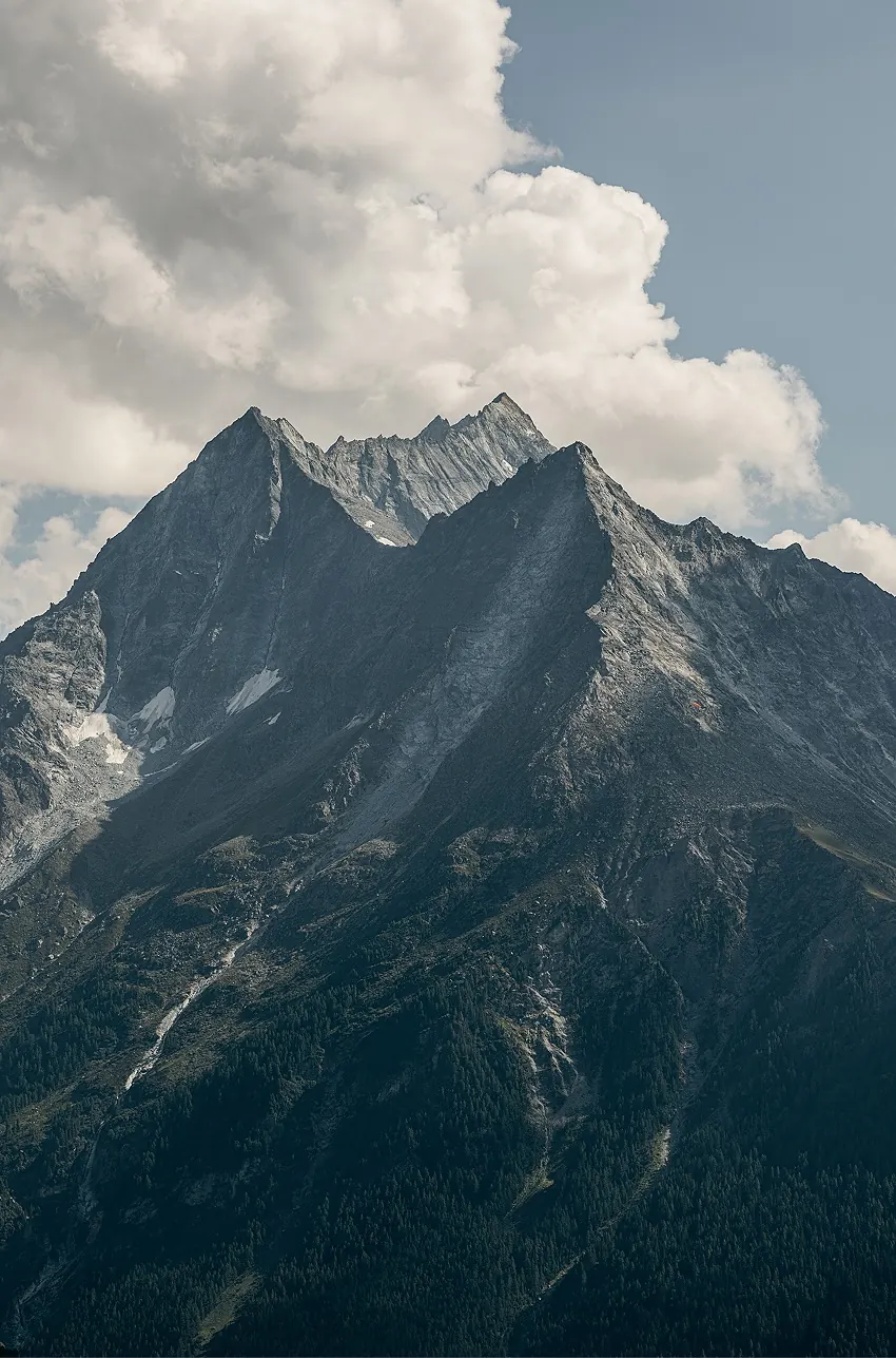 Dramatic alpine peak with jagged ridgeline and residual snow patches rising above pine forest under a cloudy sky