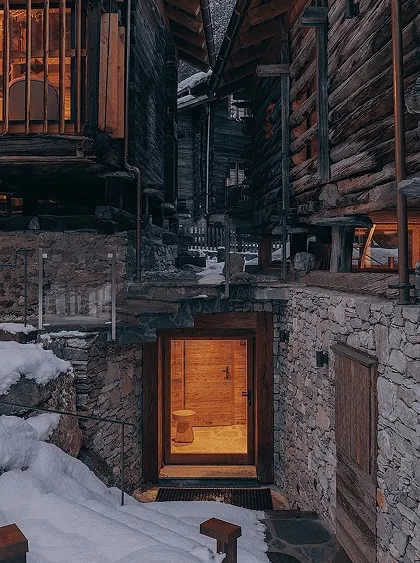 Glowing doorway cut into a stone foundation between two dark timber chalets, snow-covered village alley at dusk