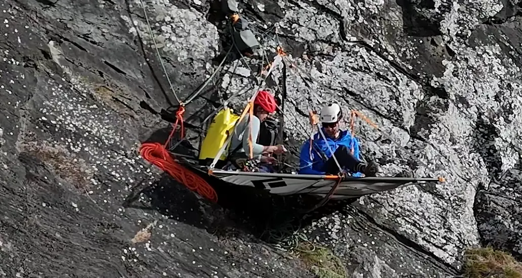 Two climbers in helmets resting on a portaledge fixed to a granite rock face, ropes and gear visible