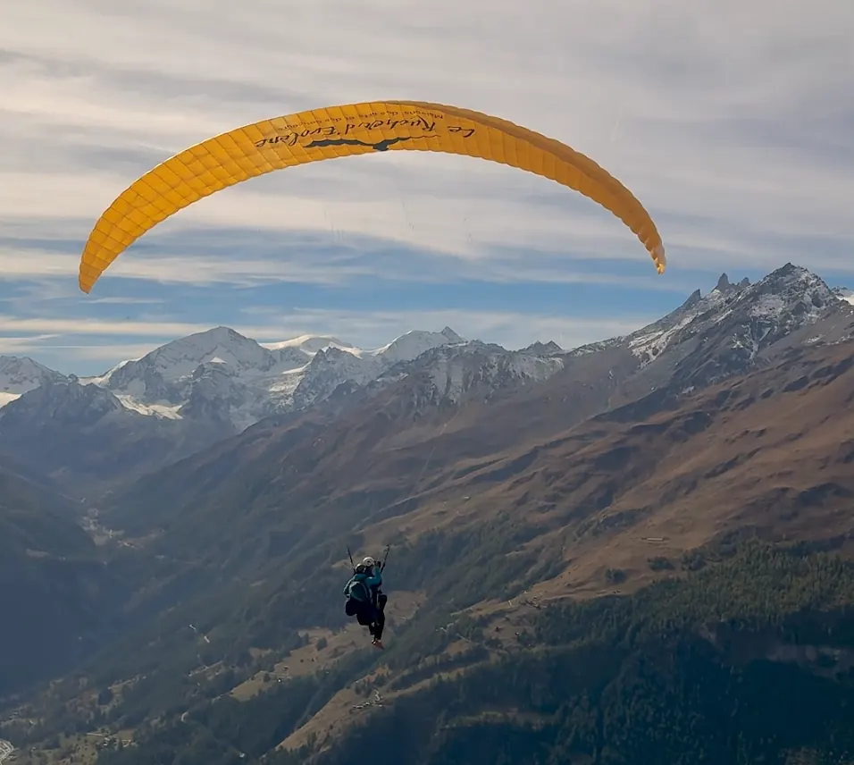 Paraglider with a yellow canopy soaring above the Évolène valley, glaciated peaks stretching to the horizon
